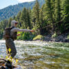 A person wearing fishing gear casts a line into a flowing river surrounded by trees and mountains on a sunny day, capturing the spirit of Sun Valley Fly Fishing in Idaho.