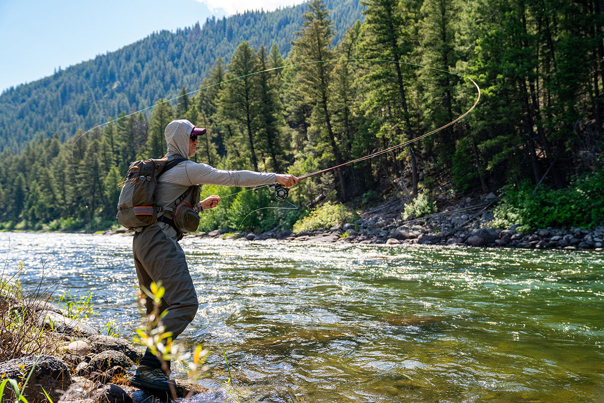 A person wearing fishing gear casts a line into a flowing river surrounded by trees and mountains on a sunny day, capturing the spirit of Sun Valley Fly Fishing in Idaho.