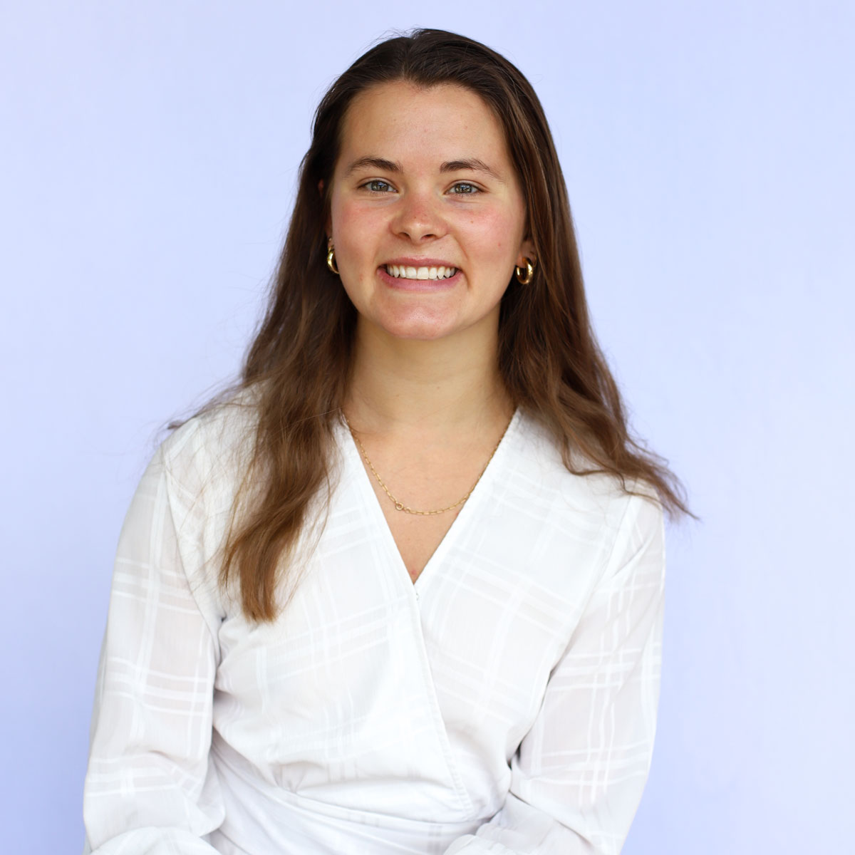 A woman with long brown hair, wearing a white blouse and gold jewelry, smiles in front of a light purple background—Melissa Hammer from Shipping and Receiving.