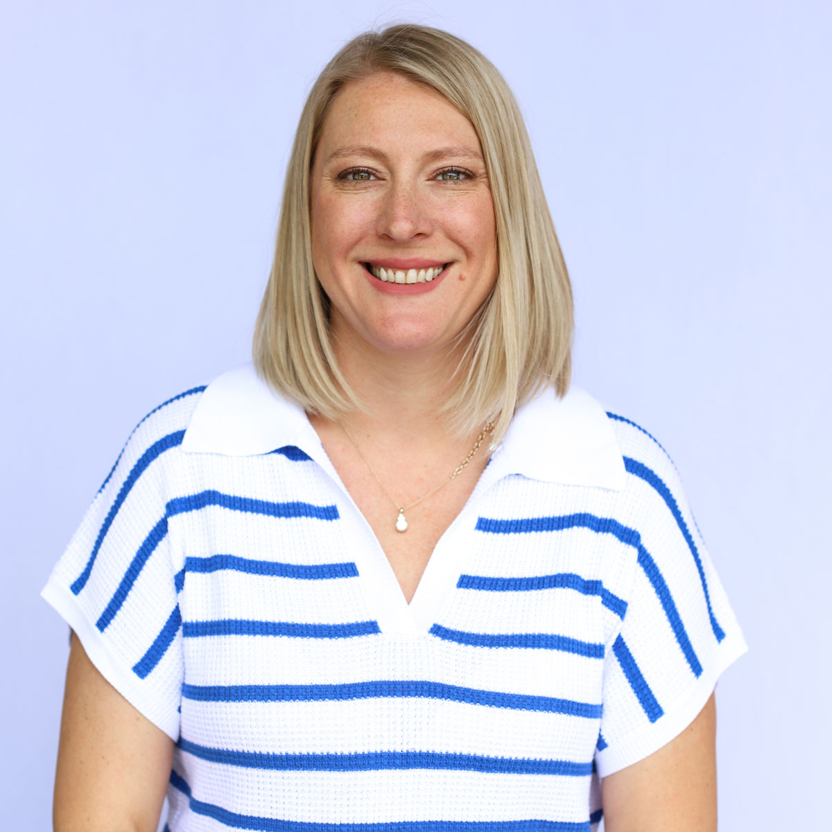 A smiling woman with straight blonde hair, who is a Buyer, wears a white shirt with blue stripes and a pendant necklace, standing in front of a light purple background.