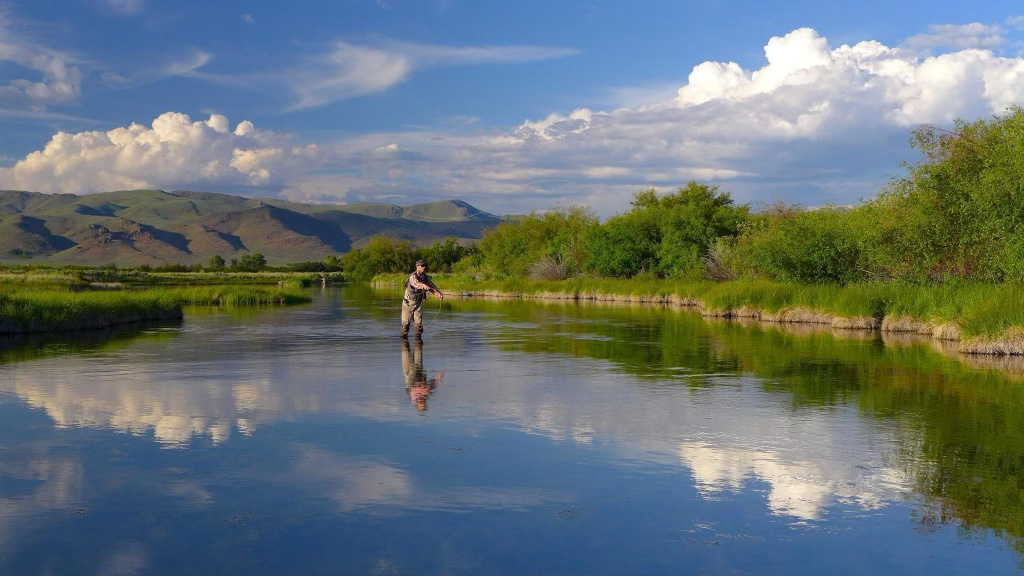A person fly fishing Idaho in a calm river surrounded by green bushes, with mountains and a blue sky with clouds reflected in the water. This picturesque setting is reminiscent of the serene views highlighted in the Fly Fishing Forecast July 8-15 | Sun Valley Idaho.