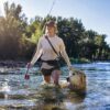 A person wearing a hat and shorts is fly fishing in a shallow river, holding a fishing rod, while a golden retriever walks beside them. The scene captures the essence of Sun Valley Fly Fishing, with trees and blue sky in the background enhancing the peaceful atmosphere.