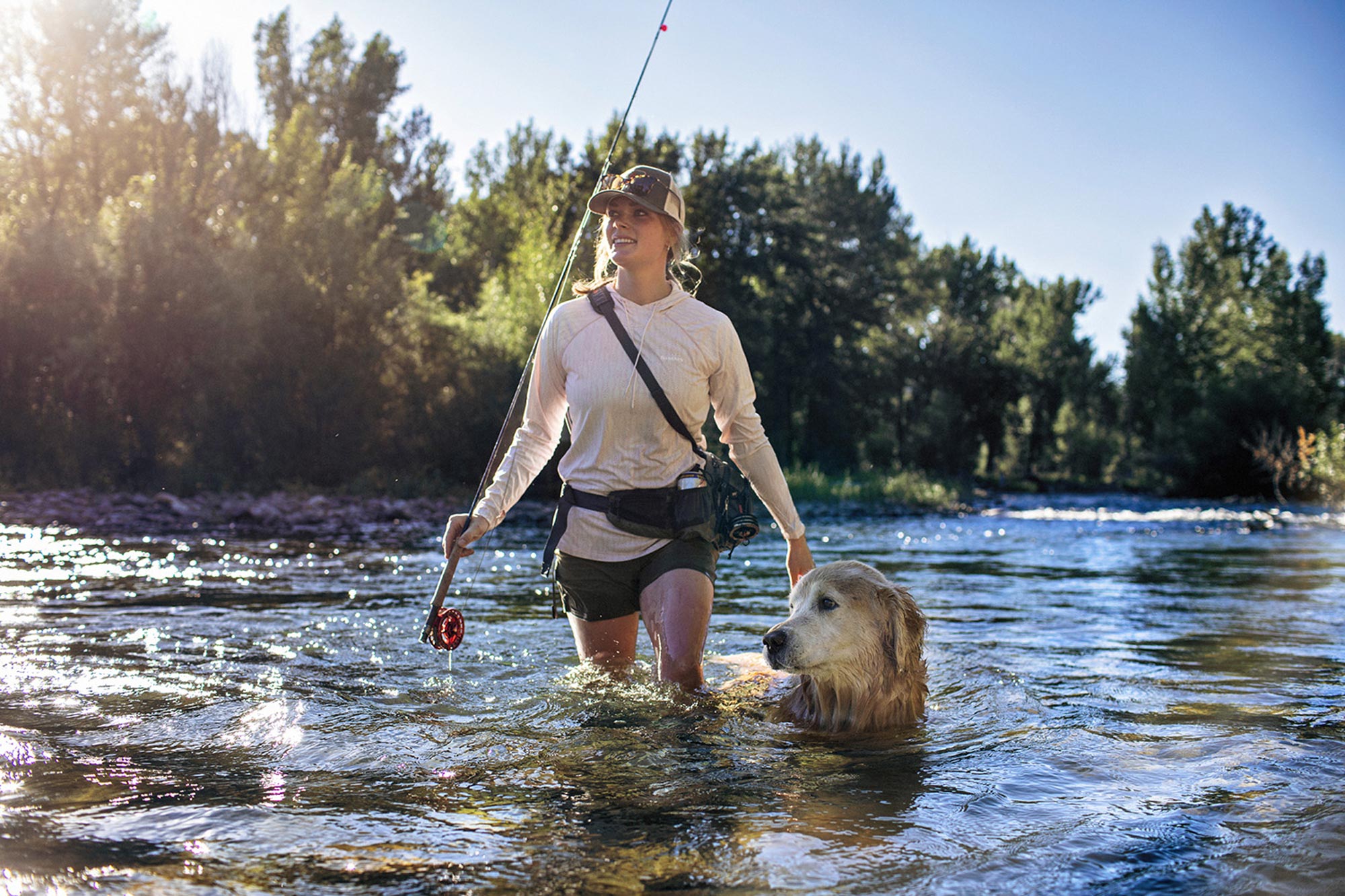 A person wearing a hat and shorts is fly fishing in a shallow river, holding a fishing rod, while a golden retriever walks beside them. The scene captures the essence of Sun Valley Fly Fishing, with trees and blue sky in the background enhancing the peaceful atmosphere.