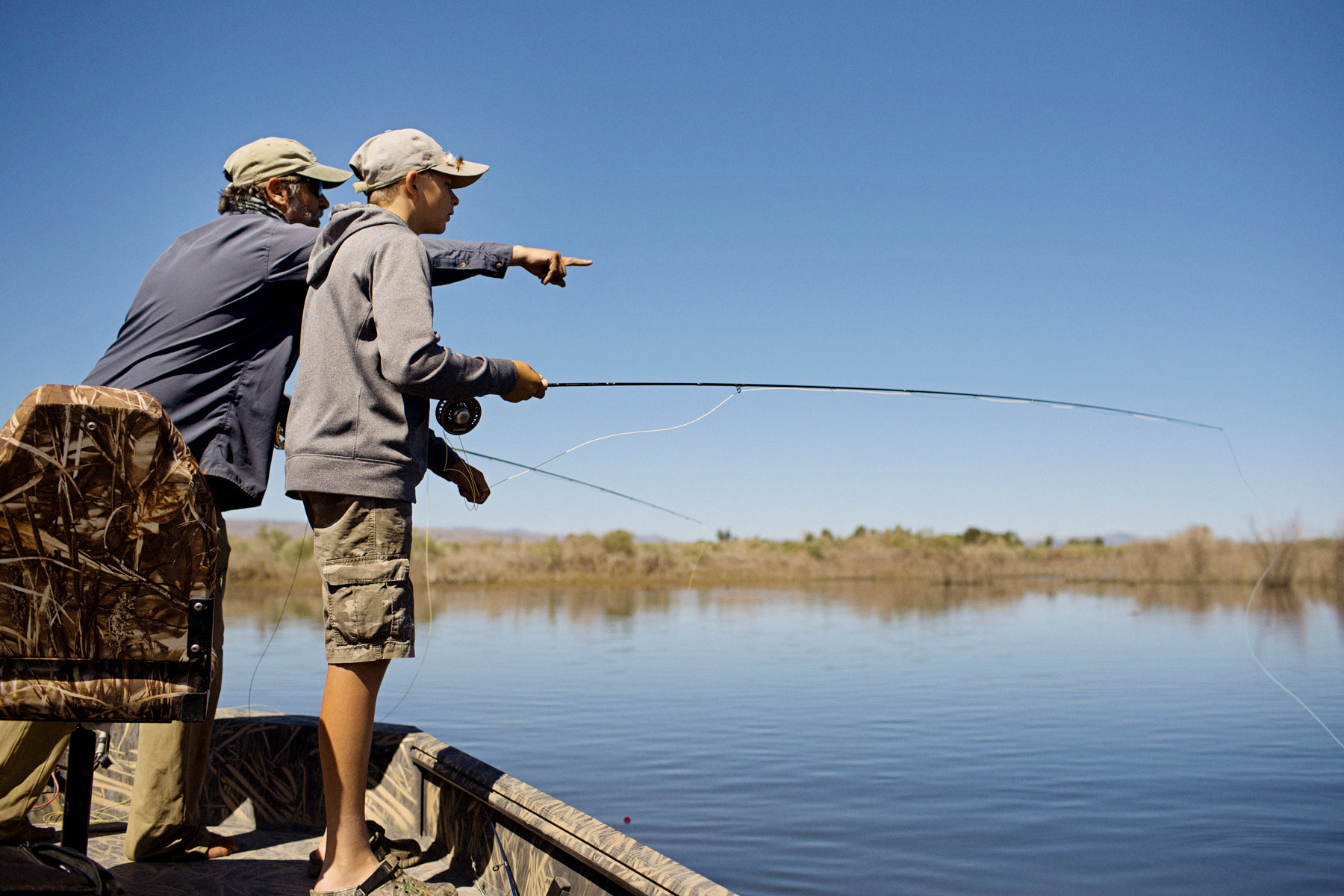 Two people in casual clothes stand on a boat, holding a fishing rod, embodying the spirit of Sun Valley Fly Fishing. One points toward the water under a clear blue sky as they discuss their Fly Fishing Forecast for August 18-19. Reeds and trees are visible in the distance, setting a tranquil scene typical of Sun Valley, Idaho.