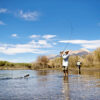 Two people stand in a shallow river, winter fly fishing Idaho under a blue sky with mountains in the background. One person is reeling in a fish as the other stands farther back in the water, making the most of ideal conditions highlighted in the Fly Fishing Forecast August 12-18 | Sun Valley Idaho.
