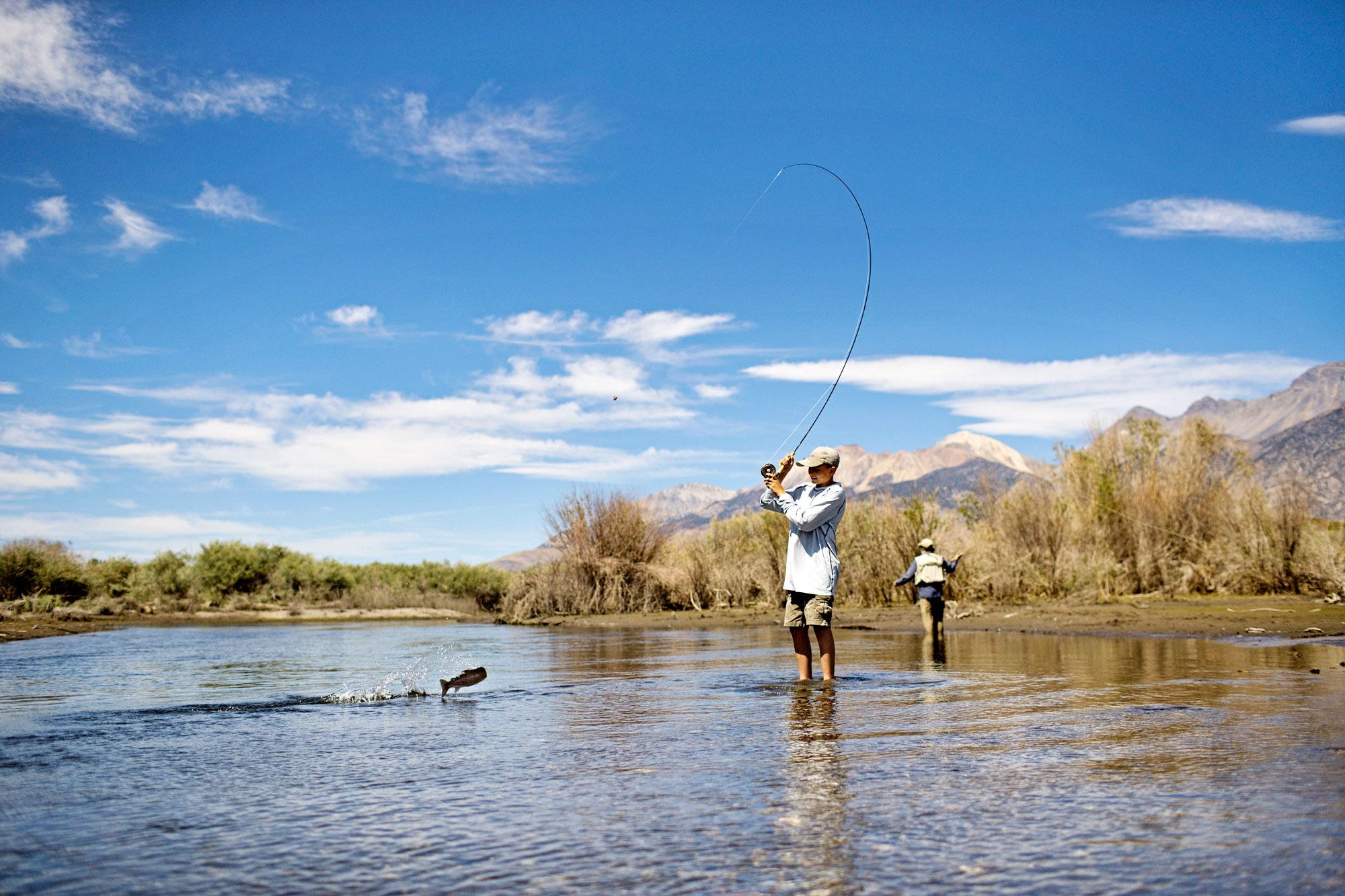 Two people stand in a shallow river, winter fly fishing Idaho under a blue sky with mountains in the background. One person is reeling in a fish as the other stands farther back in the water, making the most of ideal conditions highlighted in the Fly Fishing Forecast August 12-18 | Sun Valley Idaho.