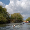 A person standing in a river uses a fishing rod to catch a fish, which is being lifted out of the water with a net. Bushes and a cloudy sky are in the background, capturing the essence of fall fly fishing in Idaho. Silver Creek Outfitters