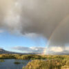 A faint rainbow appears under cloudy skies over a marsh with tall grasses and distant mountains, with sunlight breaking through the clouds—a serene scene that perfectly captures the spirit of fly fishing Idaho enthusiasts seek.