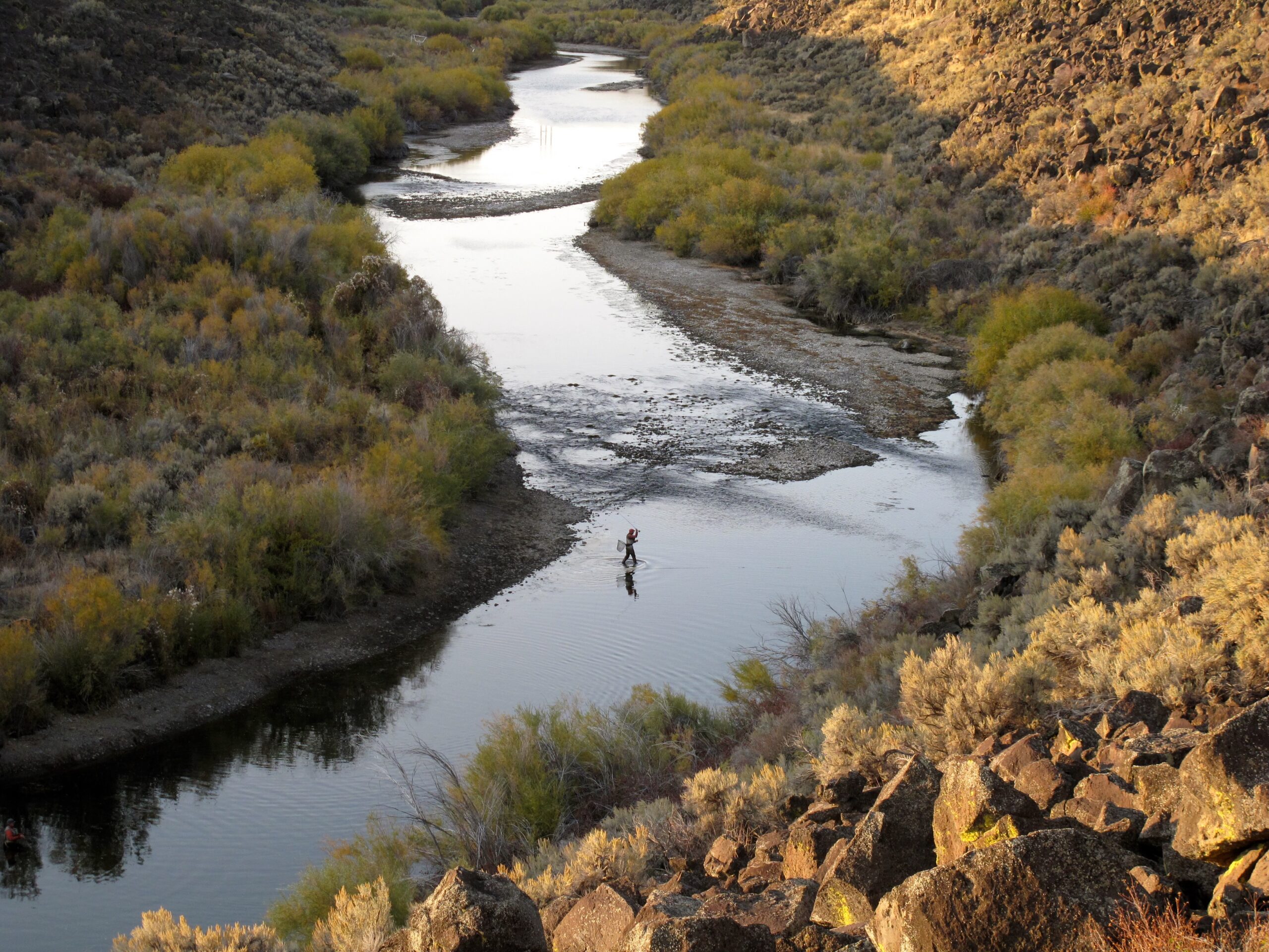A person fly fishing in a winding river canyon during a Sun Valley winter, surrounded by brush and rocky hillsides.